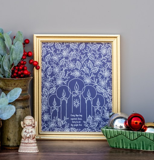The Come Thou Long Expected Jesus advent art print surrounded by a festive poinsettia floral with five advent candles, against a midnight blue background, displayed in a gold frame alongside fresh greens and berries, a ceramic bowl of ornaments, and figurine.