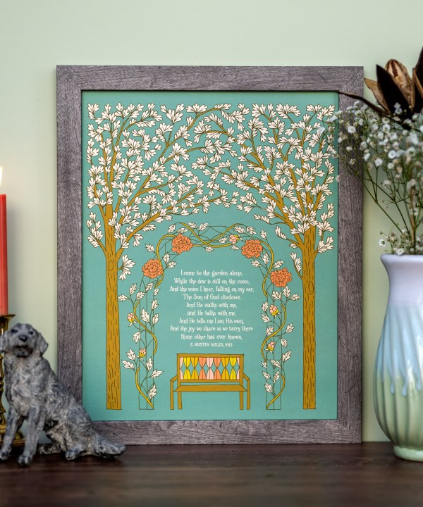 In the Garden gospel song art print features the hand lettered song text surrounded by illustration of a beautiful garden setting against a teal background, displayed in a gray wood frame, styled with a vase of flowers and greens, a candle, and a dog figurine.