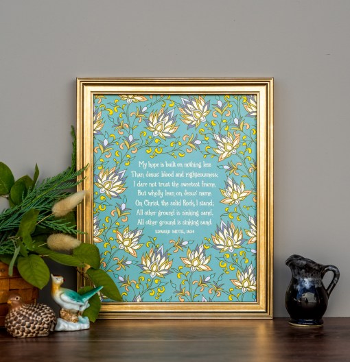 My Hope is Built on Nothing Less hymn art print features a delicate multicolored floral illustration against a teal background, displayed in a burnished gold frame, styled with greens, a vase, and ceramic figurines.