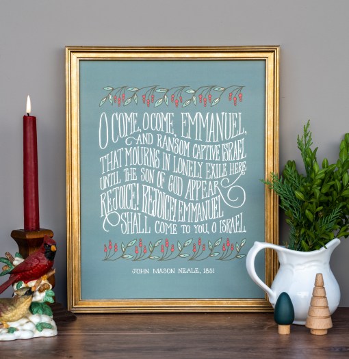 O Come O Come Emmanuel advent hymn art print, features hand-lettered text against a seasonal green background with festive leaves and berries, displayed in a gold frame, styled alongside a vase of fresh greens and a cardinal candlestick