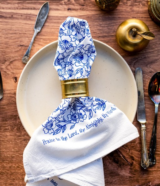 "Praise to the Lord" hymn cloth napkins use the same flour sack cotton as our tea towels. These are printed in blueberry with a beautiful floral illustration and the hymn text printed around the edge, shown styled with a napkin ring, silverware, a brass apple, and wooden table.