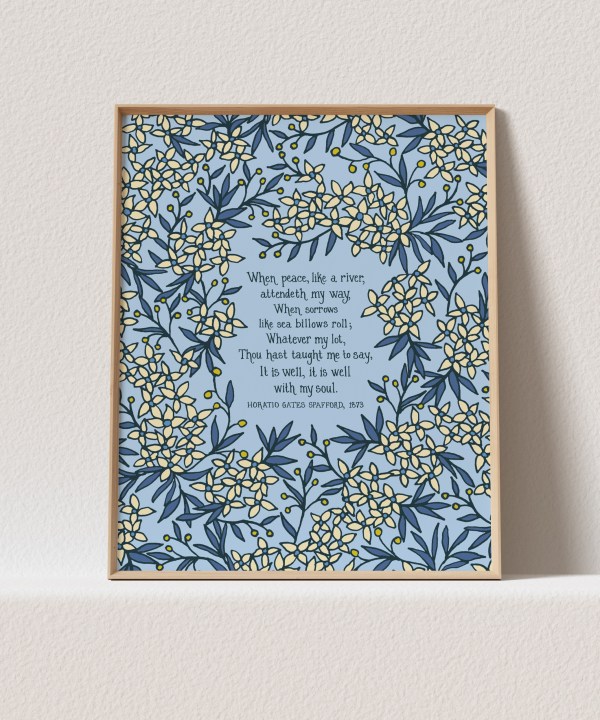 The "It Is Well" art print features the beloved hymn text surrounded by delicate floral and a sky blue background that will brighten up any room. Shown here in a light wood frame against a white background.