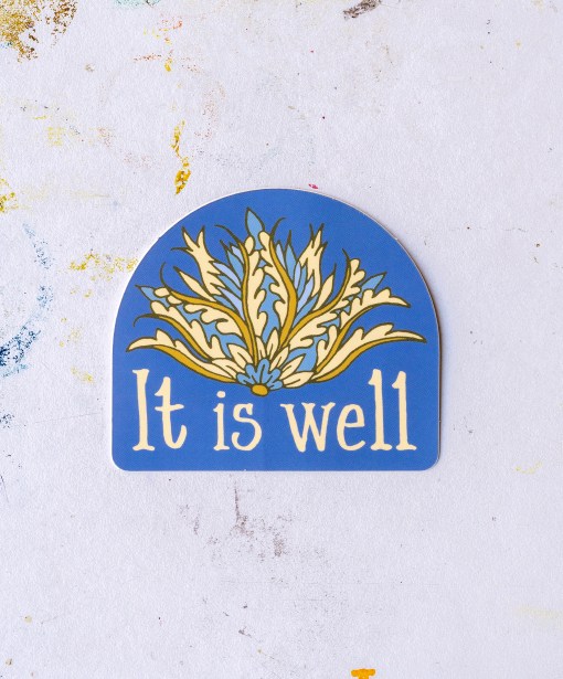 The "It Is Well" hymn sticker features an arched window shape, deep periwinkle background and floral motif in shades of blue, gold, and cream, shown against a worktable background.