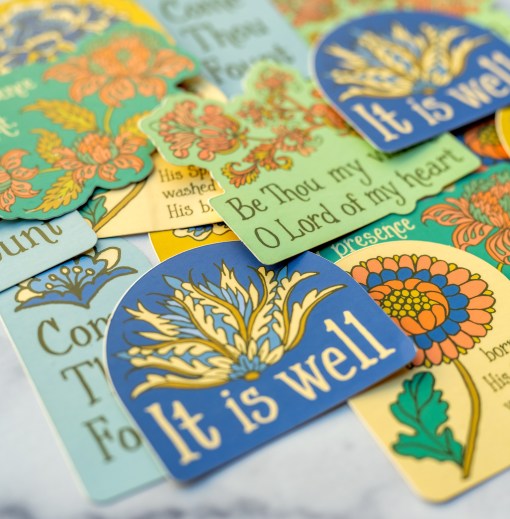 Stickers by Little Things Studio include your favorite hymn texts or quotes, accented by the beautiful illustrations and vibrant colors characteristic of Little Things Studio. Shown here as a group arrayed across a white marble background.