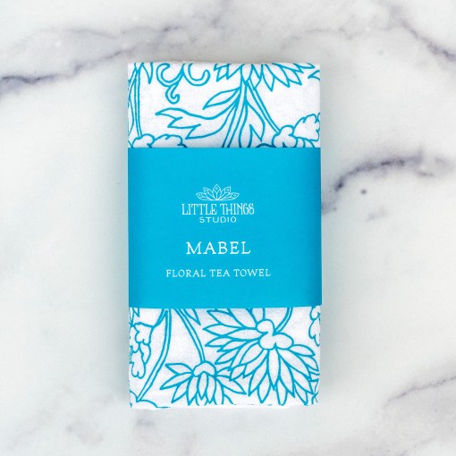 Little Things Studio presents floral tea towels — the Mabel floral tea towel features lively floral illustrations in vibrant turquoise, shown folded with a paper belly band for gift giving.