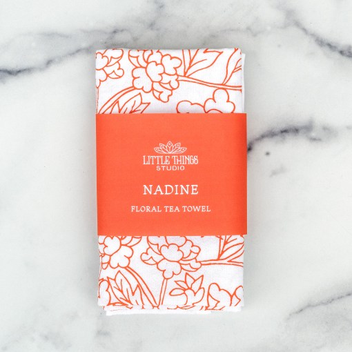 Little Things Studio presents the Nadine floral tea towel—100% cotton, made in the USA, with subtle florals in a cheerful coral, shown folded with a paper belly band for gift giving.