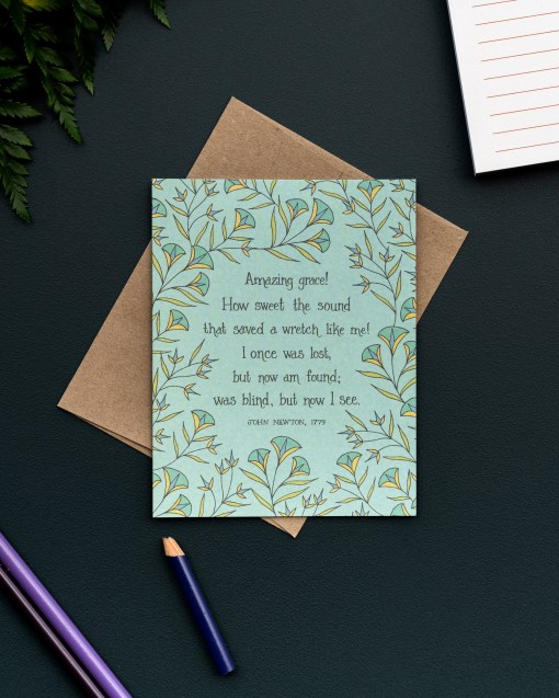 The Amazing Grace hymn greeting card, framed by dark mint and vintage yellow floral against a baby green background, is styled with a kraft paper envelope, colored pencils, notepad, and fresh greens.