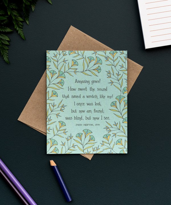 The Amazing Grace hymn greeting card, framed by dark mint and vintage yellow floral against a baby green background, is styled with a kraft paper envelope, colored pencils, notepad, and fresh greens.