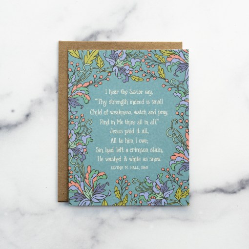 The Jesus Paid It All hymn greeting card, framed by grapefruit and asparagus colored floral against a blue lagoon background, shown with a kraft paper envelope against a marble background.