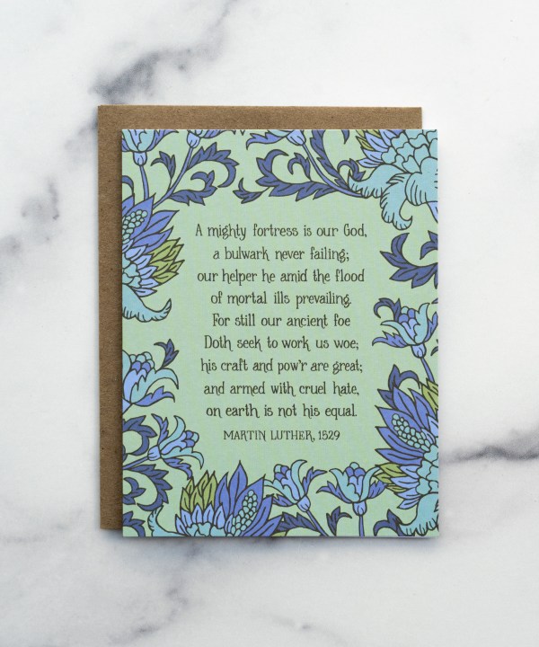 The A Mighty Fortress greeting card, framed by thunder blue and fern floral against a pastel green background, shown with a kraft paper envelope against a marble background.