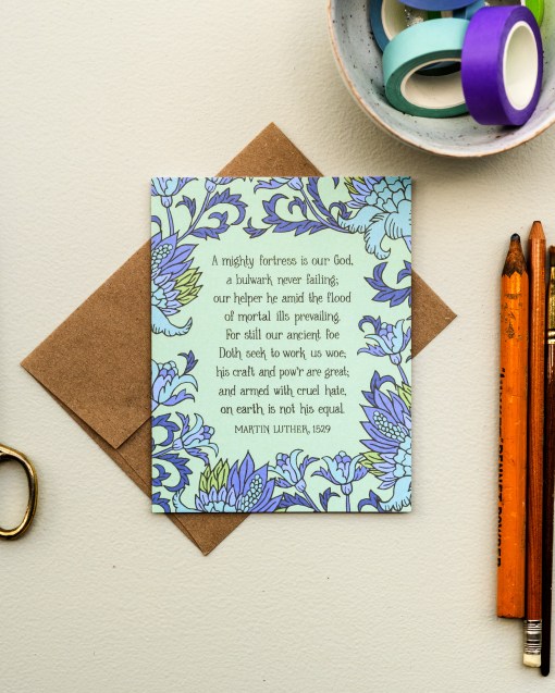 The A Mighty Fortress greeting card, framed by thunder blue and fern floral against a pastel green background, is styled with a kraft paper envelope, colored pencils, scissors, and washi tape.