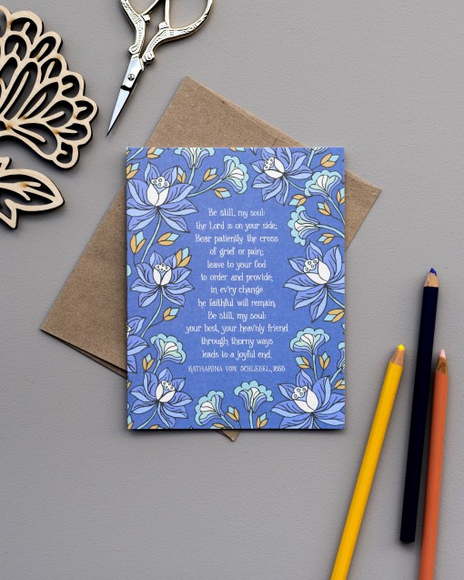 The Be Still My Soul greeting card, framed by blue violet and moonstone floral against a thunder blue background, is styled with a kraft paper envelope, colored pencils, a wooden flower stem, and scissors.