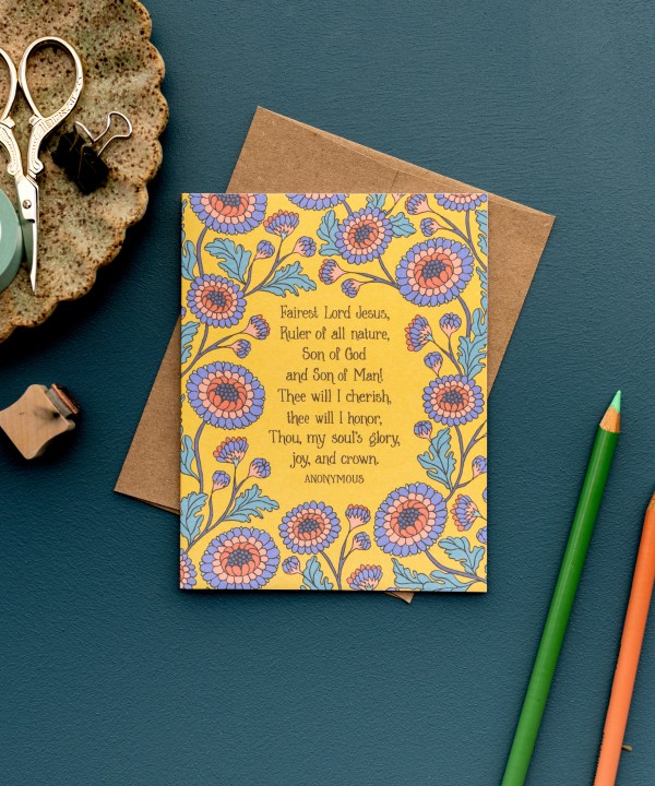 The Fairest Lord Jesus hymn greeting card, framed by blue violet and burnt sienna florals against a sunlight background, is styled with a kraft paper envelope, colored pencils, and a ceramic dish with scissors and clips.