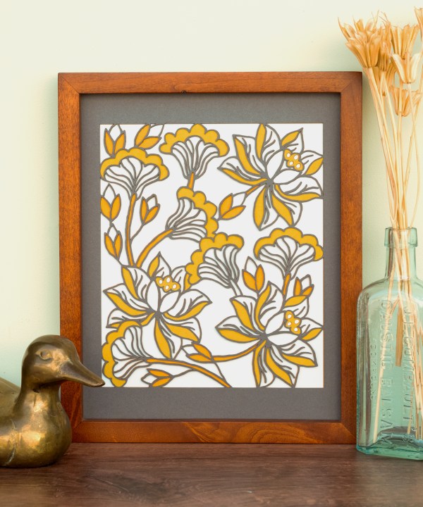 The Layered Floral Papercut “Audrey” (Light Gray) is made using cardstock in hues of dark gray, orange fizz, mustard, and light gray, shown framed and styled with a glass vase holding dried grasses and a bronze duck figurine.