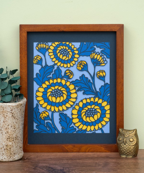 The Layered Floral Papercut “Lou” (Cornflower) is made using cardstock in hues of flag blue, Prussian blue, light gold, cornflower. Shown here framed and styled with a vase of greens and ceramic figurine.