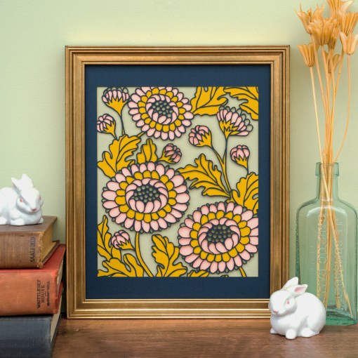The Layered Floral Papercut Lou in Kiwi is made using cardstock in hues of flag blue, mustard, salmon, and kiwi, shown here framed and styled with a vase of dried grasses, a stack of books, and two ceramic rabbits.