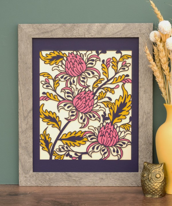 The Layered Floral Papercut “Lyra” (Violet) is made using cardstock in hues of violet, deep pretty pink, mustard, and cream, shown here framed and styled with a vase of dried flowers and a brass figurine.