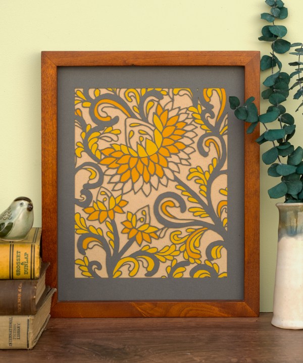 The Layered Floral Papercut “Margot” (Dark Gray) is made using cardstock in hues of dark gray, pumpkin pie, mustard, and kraft, shown framed and styled with a vase of eucalyptus and a stack of books with bird figurine.