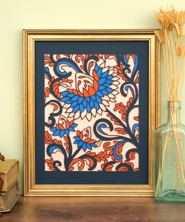 The Layered Floral Papercut “Margot” (Salmon) is made using cardstock in hues of flag blue, tangy orange, deep ocean blue, salmon, shown framed and styled with a glass vase of dried grasses, books, and a wooden figurine.