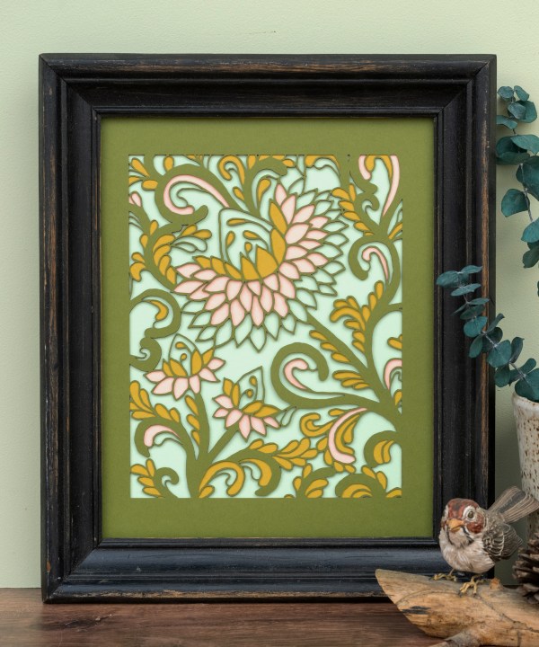 The Layered Floral Papercut “Margot” (Spearmint) is made using cardstock in hues of Jellybean green, salmon, mustard, spearmint, shown framed and styled with a vase eucalyptus, bird figurine and pinecone.