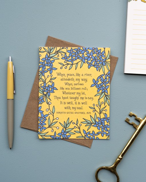The It Is Well With My Soul hymn greeting card, framed by stormy blue and lime sorbet colored florals against a buttercream background, is styled with a kraft paper envelope, ink pen, notepad, and a gold skeleton key.