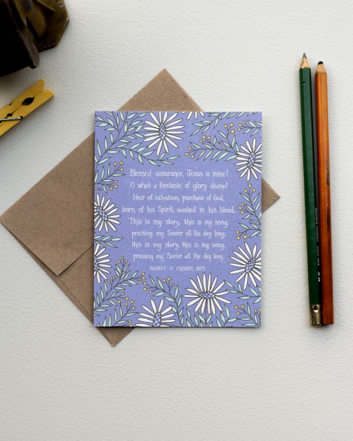 The Blessed Assurance hymn greeting card, framed by moonstone and baby green floral against a lavender gray background, is styled with a kraft paper envelope and colored pencils.
