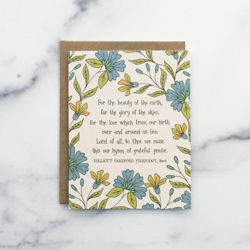 The For the Beauty of the Earth hymn greeting card, framed by asparagus and blue lagoon floral against a moonstone background, shown with a kraft paper envelope against a marble background.