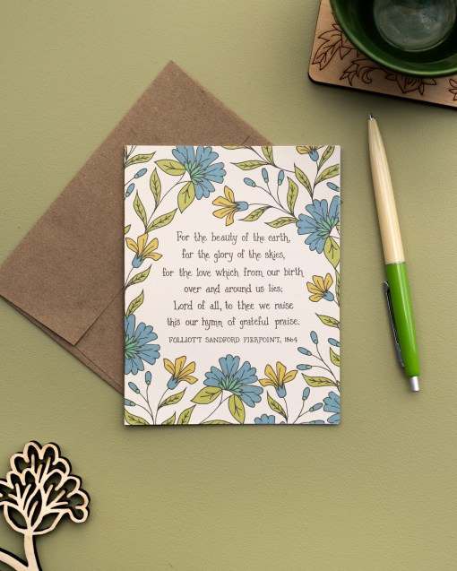 The For the Beauty of the Earth hymn greeting card, framed by asparagus and blue lagoon floral against a moonstone background, is styled with a kraft paper envelope, two-toned ink pen, and a mug with coaster.