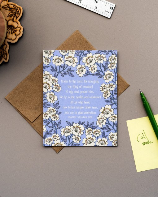 The Praise to the Lord, the Almighty hymn greeting card, framed by moonstone and lavender gray colored floral against a lilac background, is shown styled with kraft envelope, pen and sticky note.