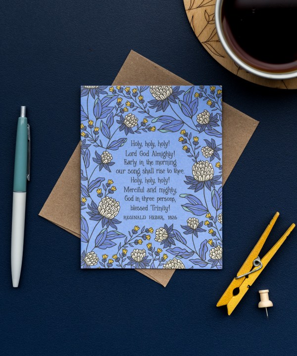 The Holy, Holy, Holy! hymn greeting card, framed by thunder blue and moonstone floral against a summer sky background, is styled with a recycled kraft paper envelope, clothes pin and thumb tack, ink pen, and a ceramic mug.