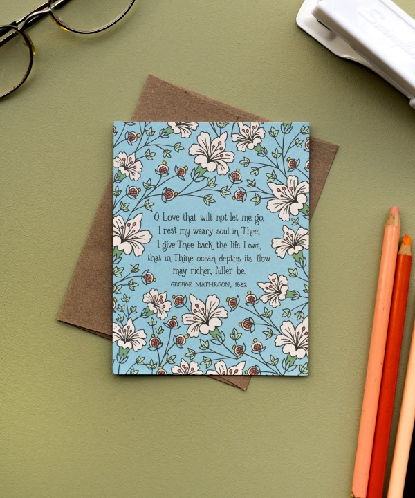 The "O Love That Wilt Not Let Me Go" hymn greeting card, framed by burnt sienna and moonstone floral against a sea mist background, styled with a recycled kraft paper envelope, colored pencils, glasses and a stapler.