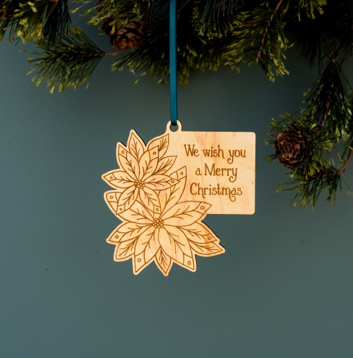 These lovely wood Christmas poinsettias ornaments from Little Things Studio usher in a festive holiday greeting filled with cheer. Engraved with florals, foliage, and lyrics from the Christmas carol “We Wish You a Merry Christmas.” Shown hanging from a tree with ribbon.