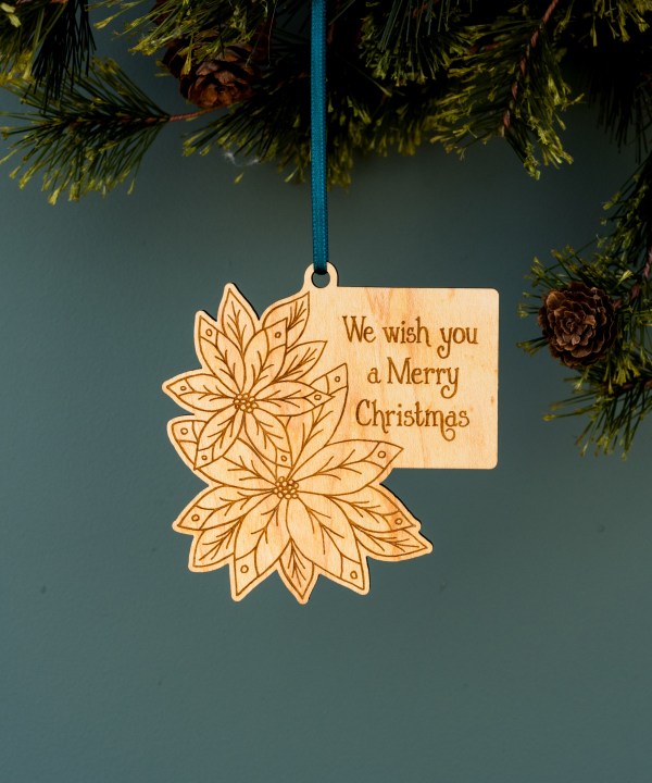 These lovely wood Christmas poinsettias ornaments from Little Things Studio usher in a festive holiday greeting filled with cheer. Engraved with florals, foliage, and lyrics from the Christmas carol “We Wish You a Merry Christmas.” Shown hanging from a tree with ribbon.