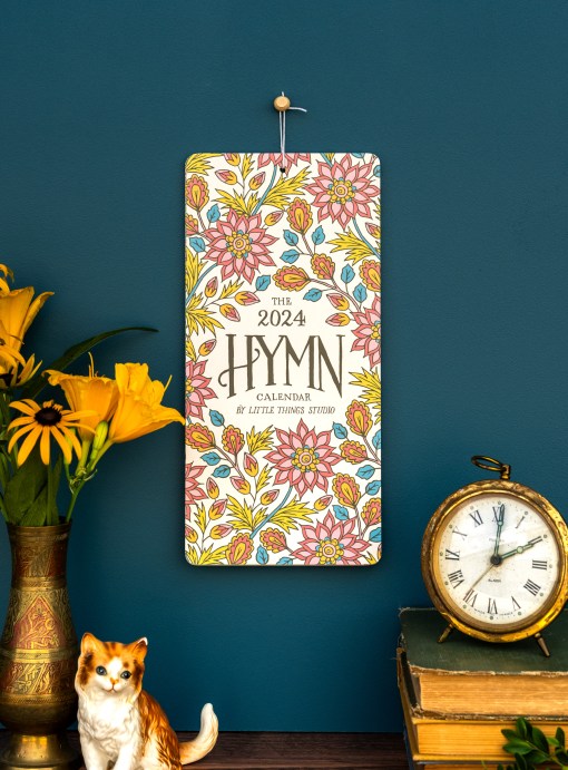 Celebrate 2024 with uplifting hymns, vibrant floral patterns, and beautiful hand-lettering, shown styled with a vase of flowers, ceramic cat, gold desk clock and books.
