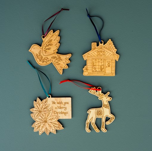 We wish you a merry Christmas with this new set of 4 ornaments by Little Things Studio, laser cut in-house from USA maple plywood with a natural oil finish, featuring Peaceful Dove, Winter House, Christmas Poinsettia, and Prancing Reindeer, shown here lying flat and tied with ribbon, ready to hang.