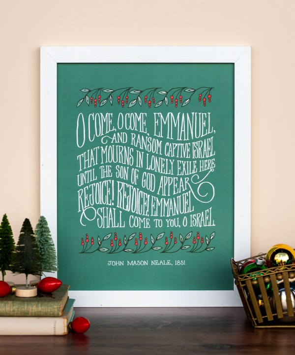 The "O Come, O Come Emmanuel" hymn art print features a verse printed in hand-lettered text against a deep green background with festive leaves and berries, shown styled in a white frame with bristle brush pine trees, books and a wooden basket of ornaments.