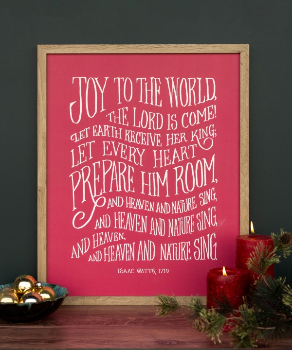 Brighten your holiday home with the "Joy to the World" hymn art print in 16" x 20", printed in hand-lettered text against a festive garnet background, shown here styled in a frame with candles and a bowl of jingle bells.