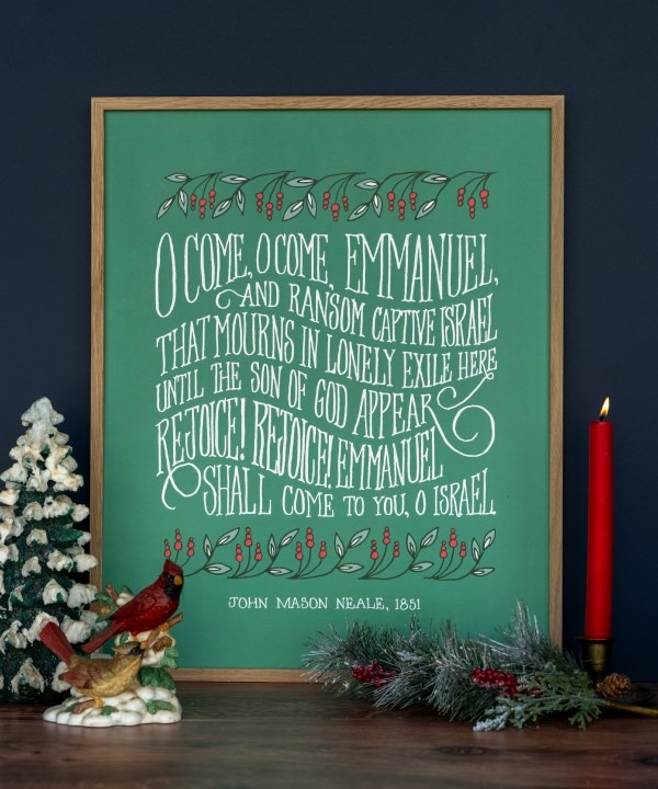 The "O Come, O Come Emmanuel" art print features a verse printed in hand-lettered text against an alpine green background with festive leaves and berries, shown here framed and styled with a candle, snowy pine tree, and cardinal bird figurine.