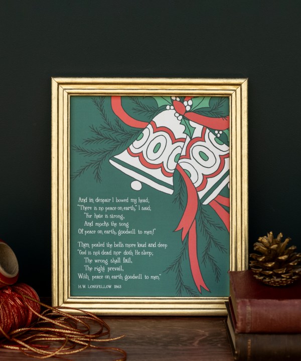 "I Heard the Bells on Christmas Day" hymn art print features a verse from the beloved seasonal hymn accented with bells against a deep green background, shown here in a gold frame styled with books, a pinecone, and festive cords.