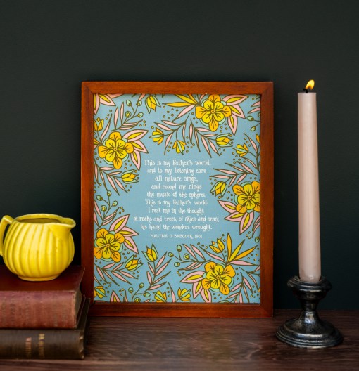 The "This is My Father's World" hymn art print features a hand-lettered verse of the beloved hymn surrounded by gorgeous floral illustration, framed and styled with books, a creamer and candlestick.